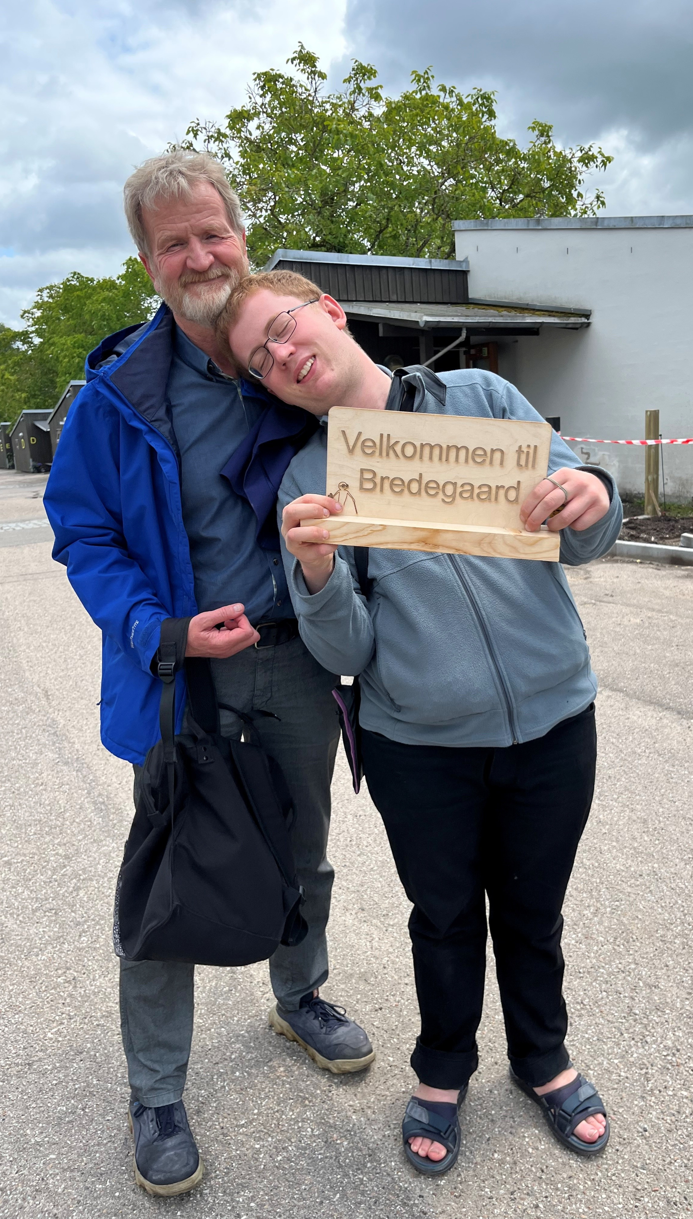 Olav sammen med sin far Niels-Bjørn. Olav holder et fint skilt i træ i sine hænder, hvor der står "Velkommen på Bredegaard."