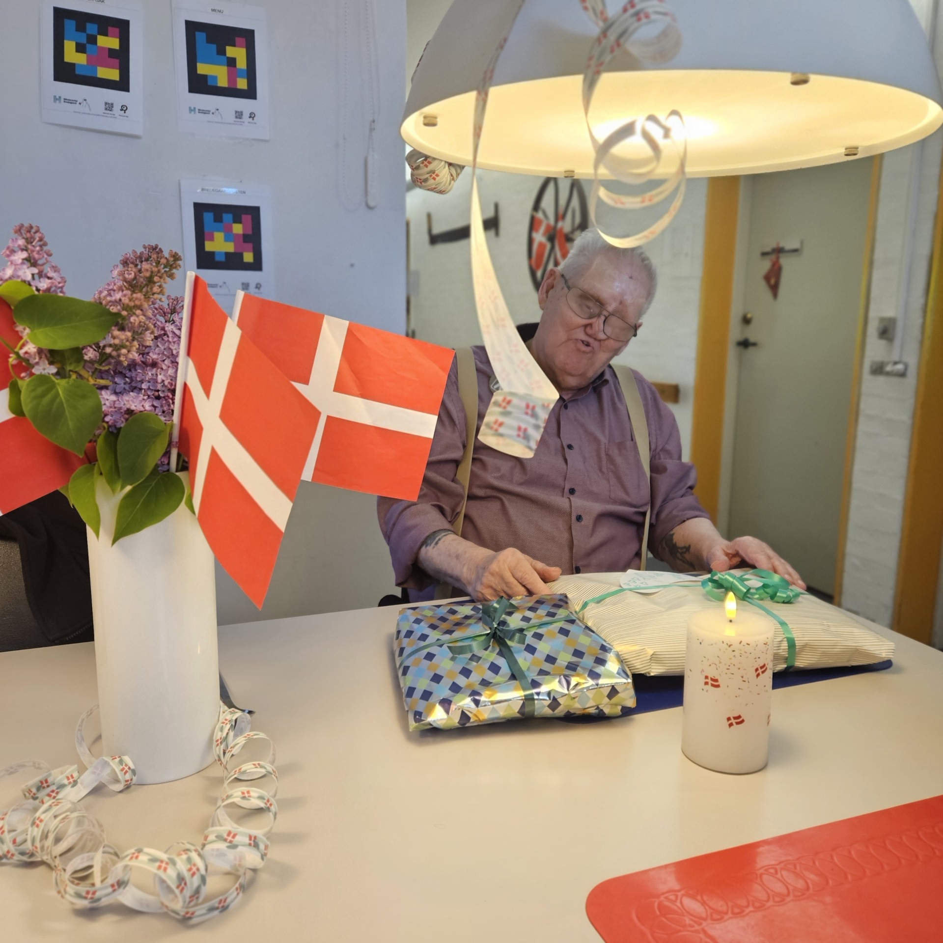 Frank Thomsen sidder ved spisesbordet i køkkenet med gaver på bordet, flag og blomster i en vase og fødselsdagspynt. 