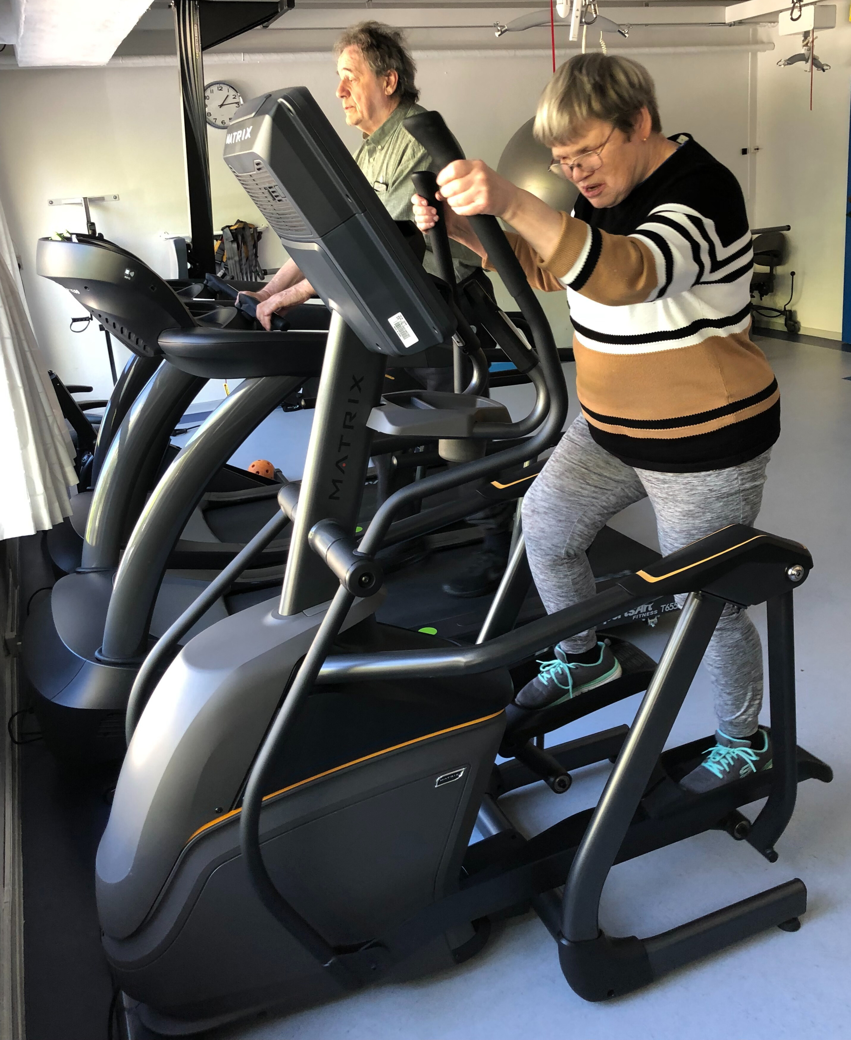 Ulla træner på den nye crosstrainer, som står i motionsrummet. Svend går på løbebånd ved siden af. 
